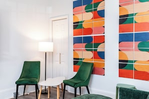 Ein Zimmer mit zwei grünen Stühlen, einem kleinen weißen Tisch und einem farbenfrohen, abstrakten Wandbild.