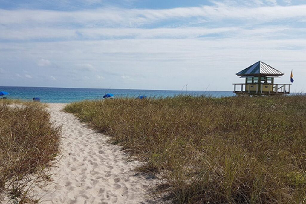 Un sentier sablonneux menant à une plage où se trouvent un poste de surveillance des sauveteurs et des parasols bleus.