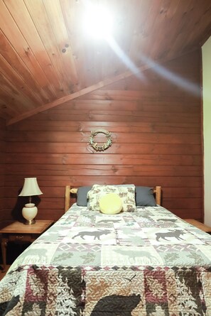 A cozy bedroom with a wooden ceiling, a bed with patterned bedding, a nightstand with a lamp, and a wall-mounted wreath.