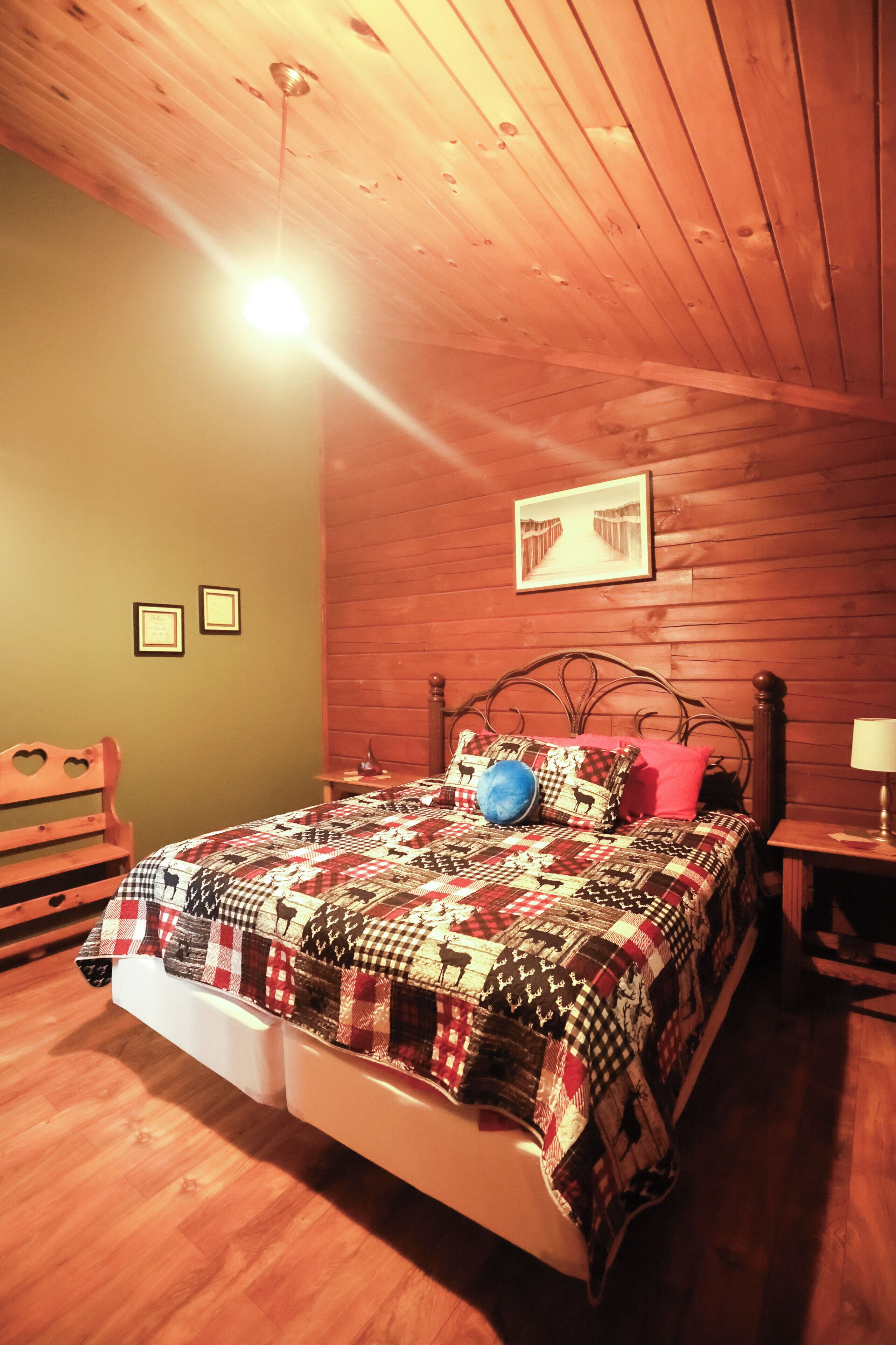 A room with a wooden ceiling, a bed with a patterned quilt, a wooden bench, and a framed picture on the wall.