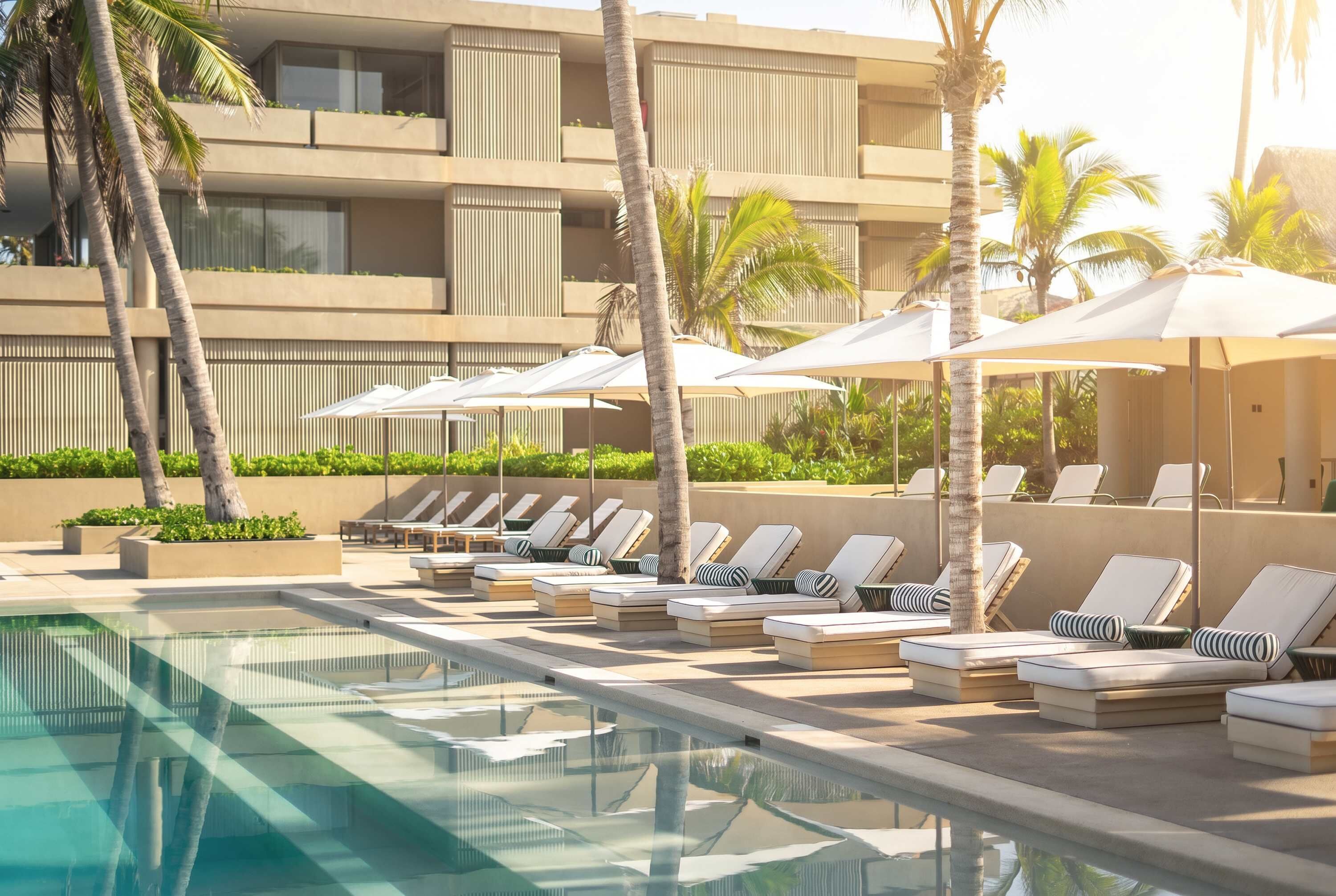 Zona de piscina con sillones de descanso y sombrillas, una piscina cristalina y un edificio con balcones.