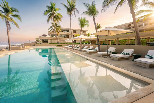A poolside area with lounge chairs and umbrellas, surrounded by palm trees and a building with multiple balconies.