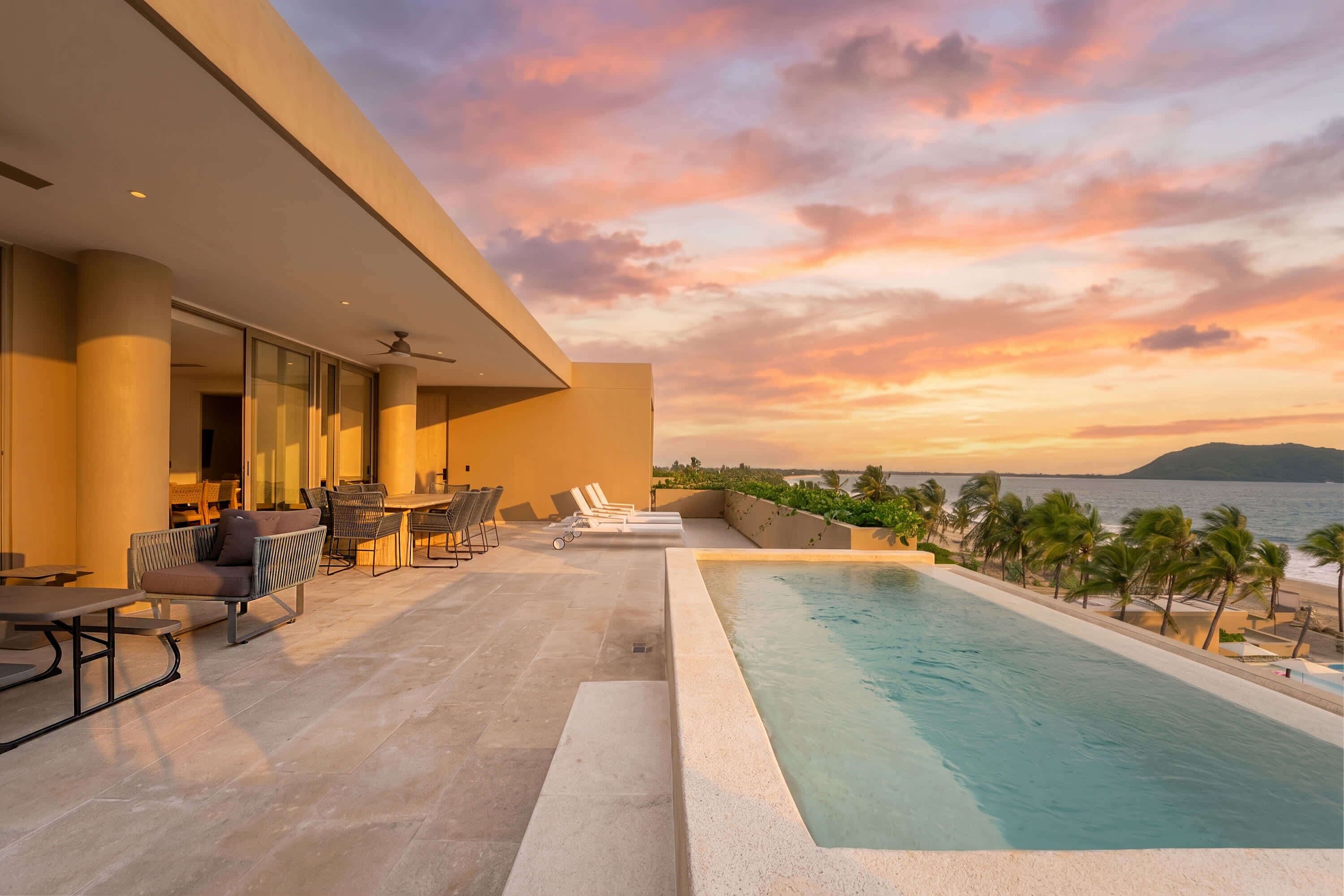 Ein Poolbereich mit Blick auf das Meer bei Sonnenuntergang, Sitzgelegenheiten im Freien und eine moderne Gebäudefassade.