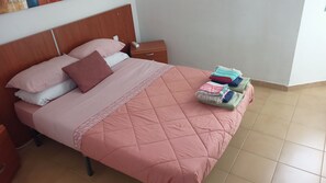 A neatly made bed with a pink quilt and pillows, a bedside table with a power outlet, and a stack of folded blankets on the floor. - Bouganville Sea & Pool View Brigit Apartment—perfectly laid out for your comfort. (Nuevo Horizonte)