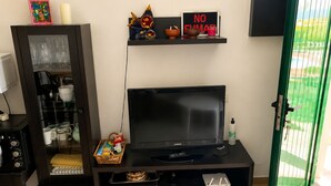 Quarto com televisão, um armário com portas de vidro, uma estante com objetos decorativos e uma porta verde.
