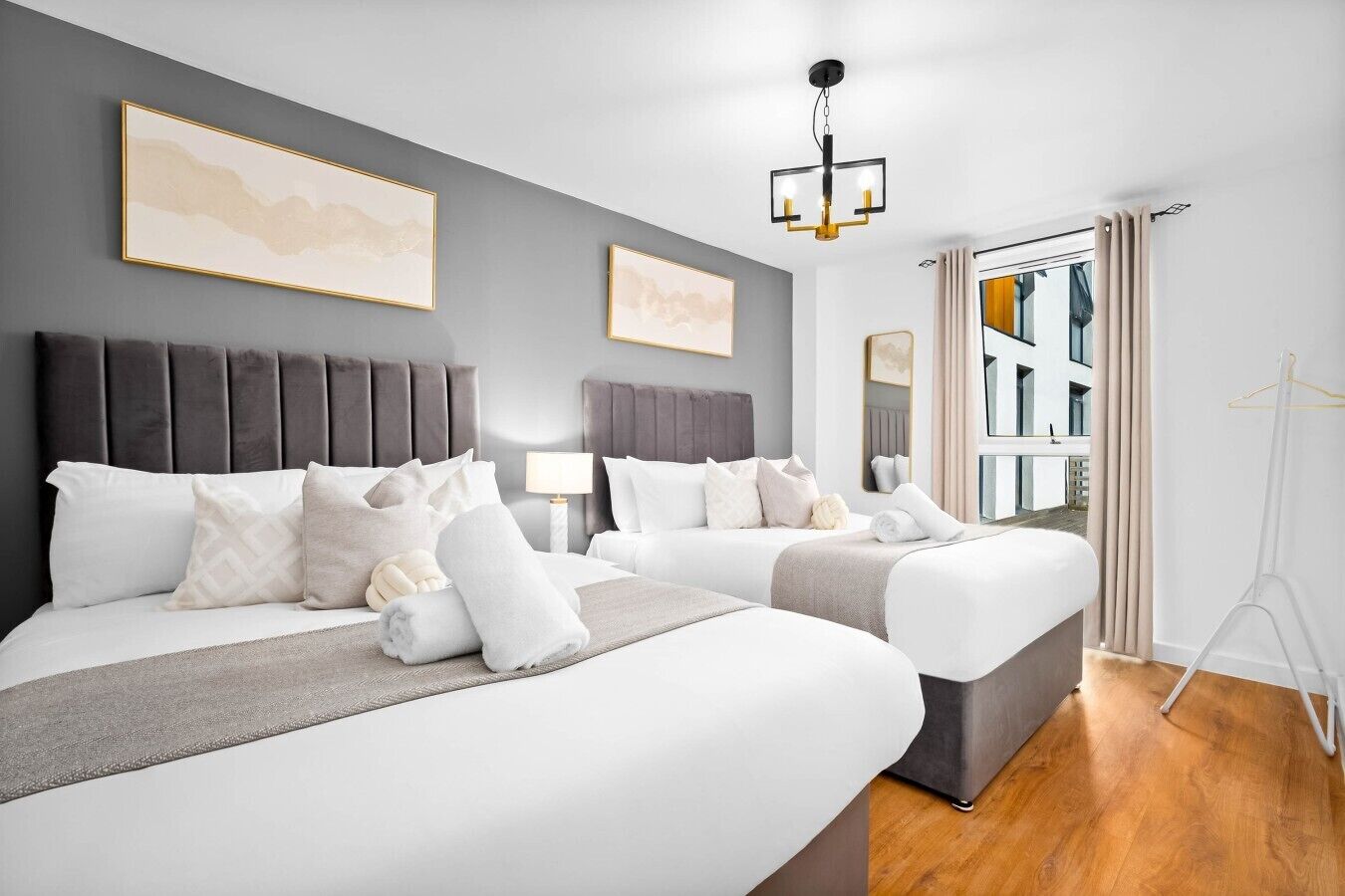 A hotel room with two beds, a grey headboard, a hanging light fixture, and a window with curtains.