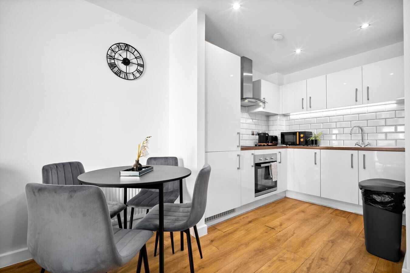 Une cuisine moderne avec des meubles blancs, une horloge noire et un coin repas avec des chaises grises.
