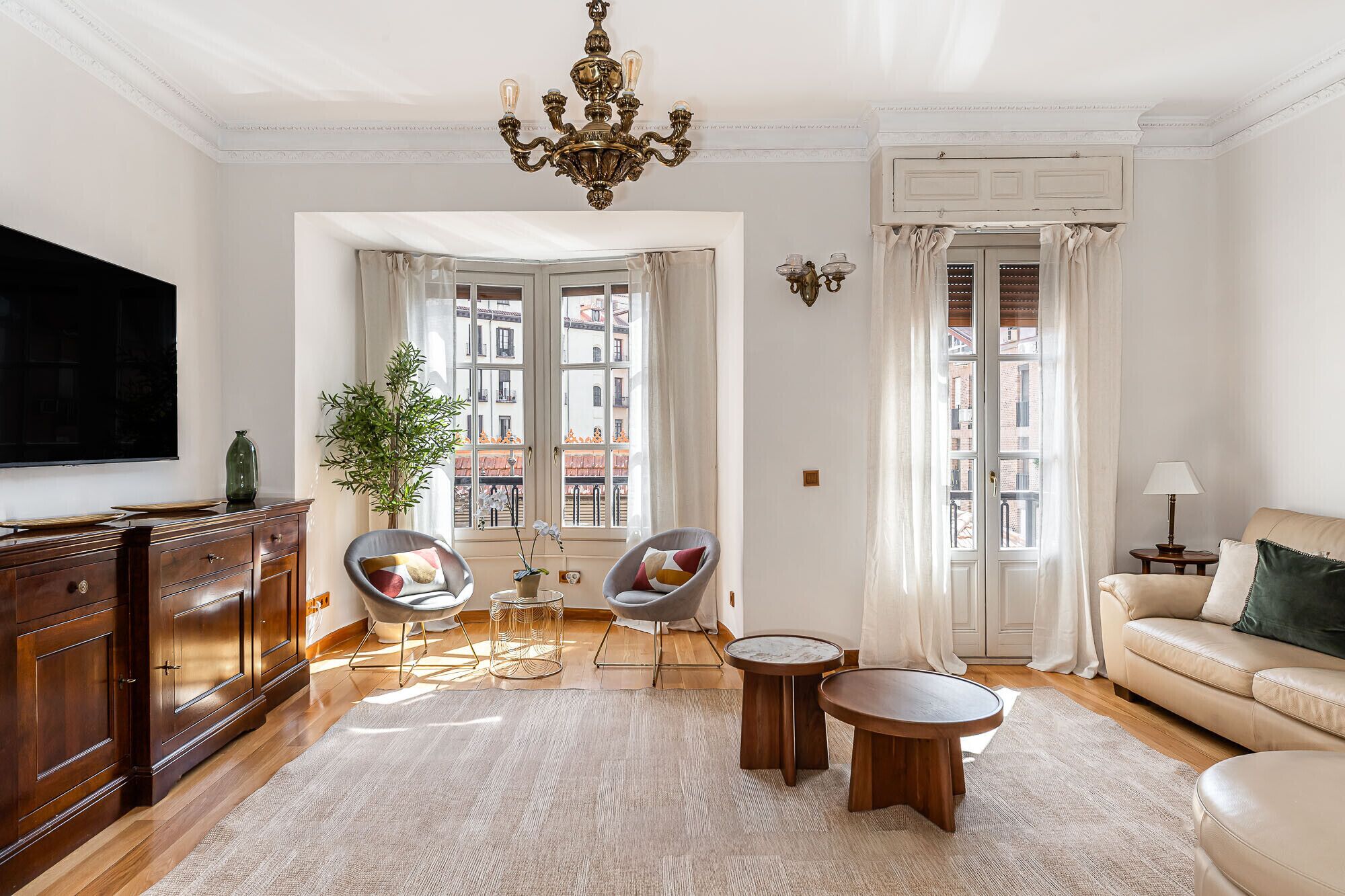 Una sala de estar con televisión, dos sillones, un sofá, una araña y vistas de la ciudad a través de amplios ventanales.