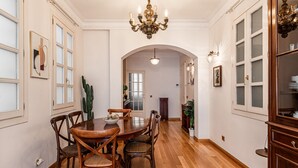 Een eetgedeelte met een houten tafel en stoelen, een kroonluchter en een doorkijkje naar een andere kamer via een boogvormige deuropening.