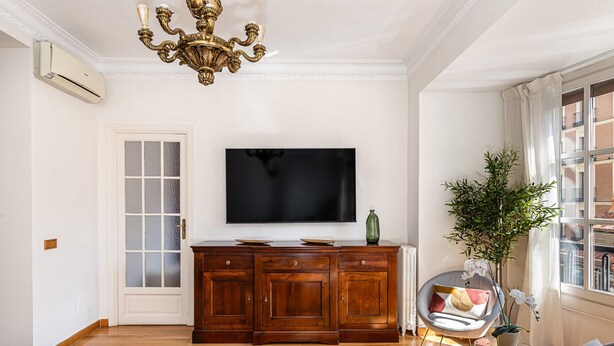 Een woonkamer met een houten kast, een flatscreen televisie, een kroonluchter, een kamerplant en een raam met uitzicht.