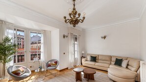 Een ruime woonkamer met een bank, twee stoelen, een kroonluchter en uitzicht op gebouwen door grote ramen.