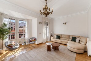 Ein geräumiges Wohnzimmer mit einer Couch, zwei Sesseln, einer Chandelieber und Blick auf Gebäude durch große Fenster.
