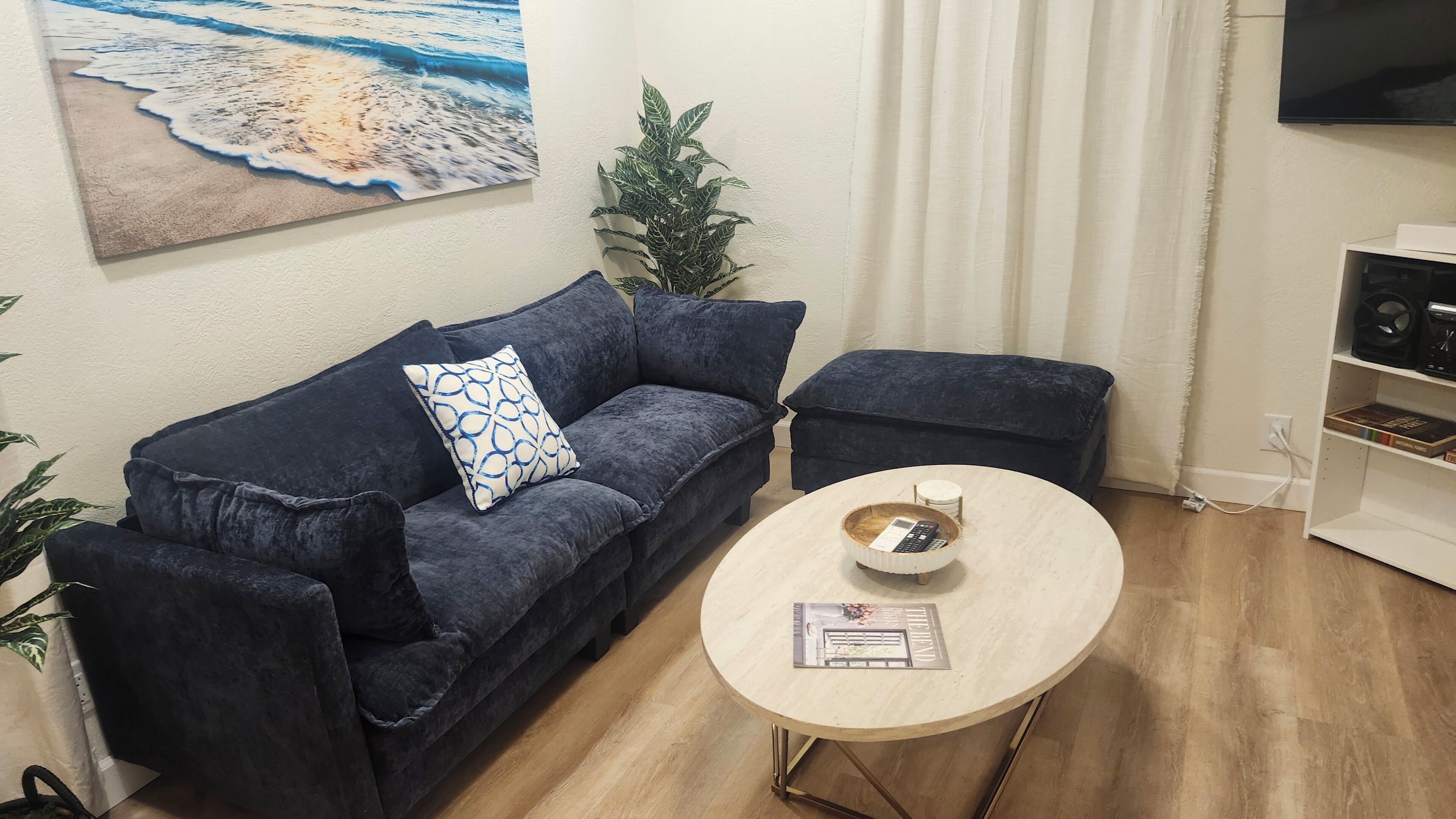 A living room with a dark blue sofa, a matching armchair, a round coffee table with a magazine, and a painting of a beach scene on the wall.
