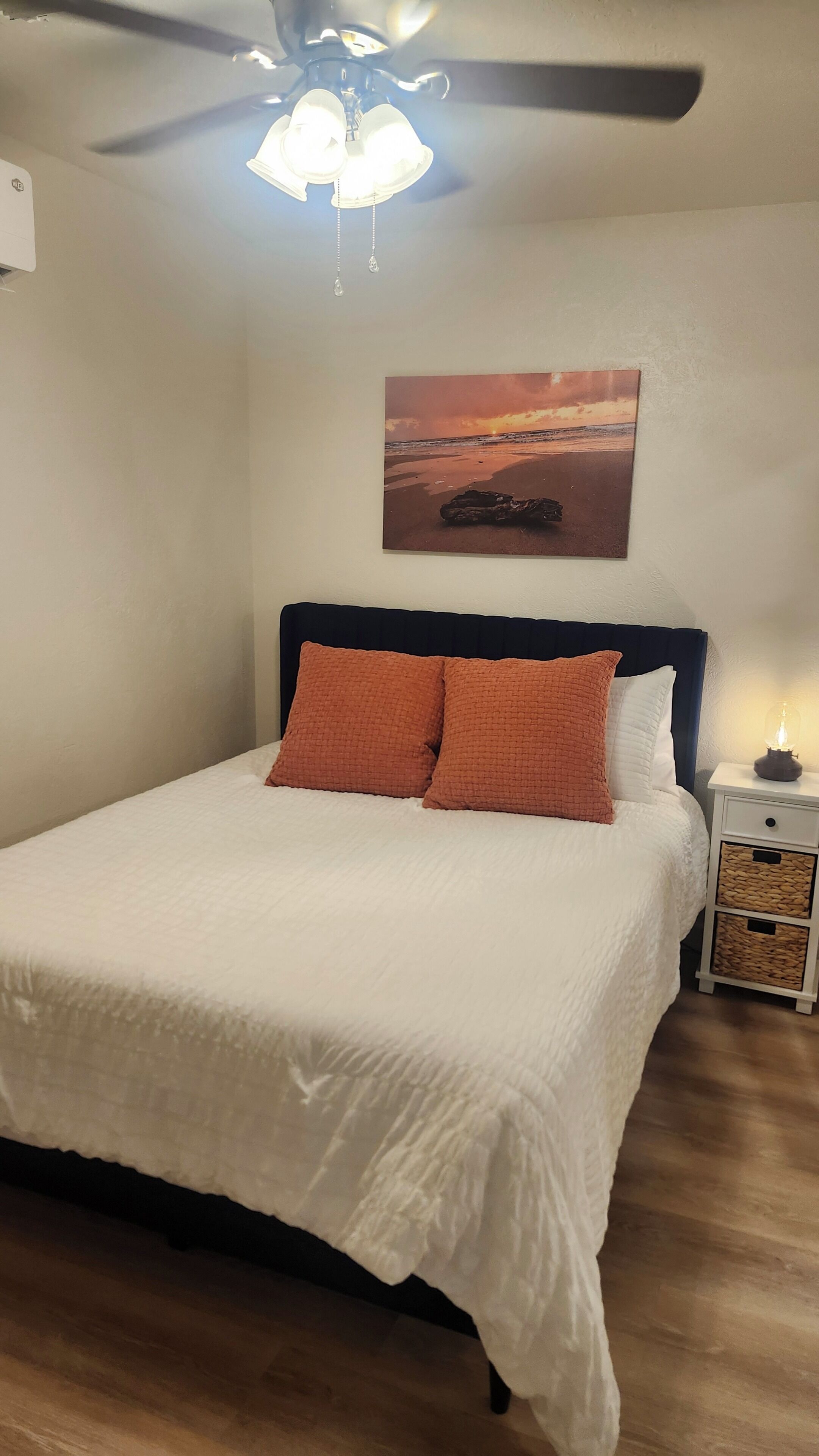 A bedroom with a bed, a ceiling fan, a wall-mounted light, a nightstand, and a picture of a sunset.