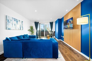 A modern living room with a blue sectional sofa, a coffee table, and a TV mounted on a wooden wall. - Luxe Two Bed Apartment - City Centre - Balcony & Parking (Birmingham)