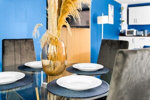Taula de menjador amb plats i un vas amb plantes seques, amb un mur blau i una cuina al fons.