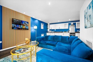 Un saló modern amb un sofà blau, una taula de centre de vidre i una televisió que mostra un partit.
