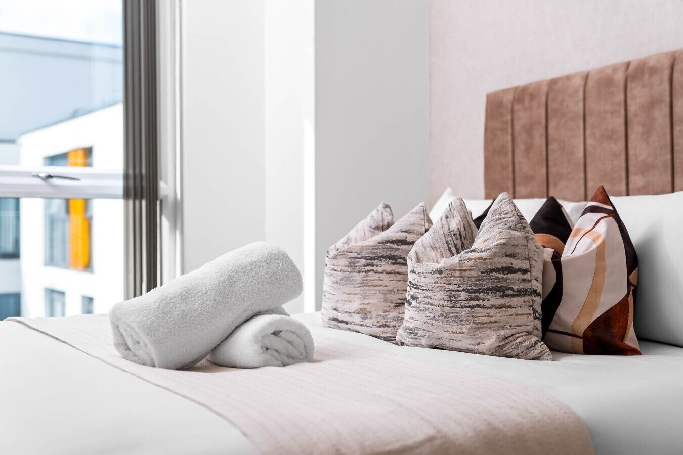 A neatly made bed with a rolled white towel, two textured pillows, and a striped pillow on a white bedspread.