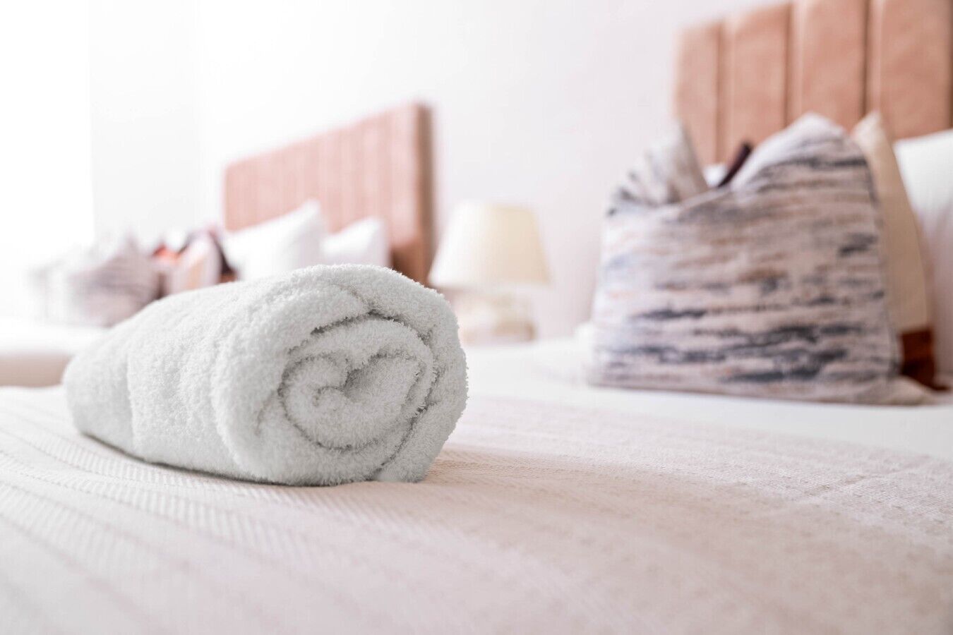 A neatly rolled white towel on a bed with a headboard and a patterned pillow in a hotel room.