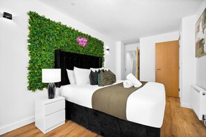 A modern bedroom with a black headboard, white bedding, a green wall-mounted plant, a neon sign, and a white bedside table. - Stylish Apartment* Jewellery Q * Parking* Netflix (West Midlands)