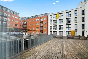 Una terraza de madera con una cerca de metal, adyacente a un edificio de ladrillo rojo con balcones y un edificio blanco con una franja amarilla.