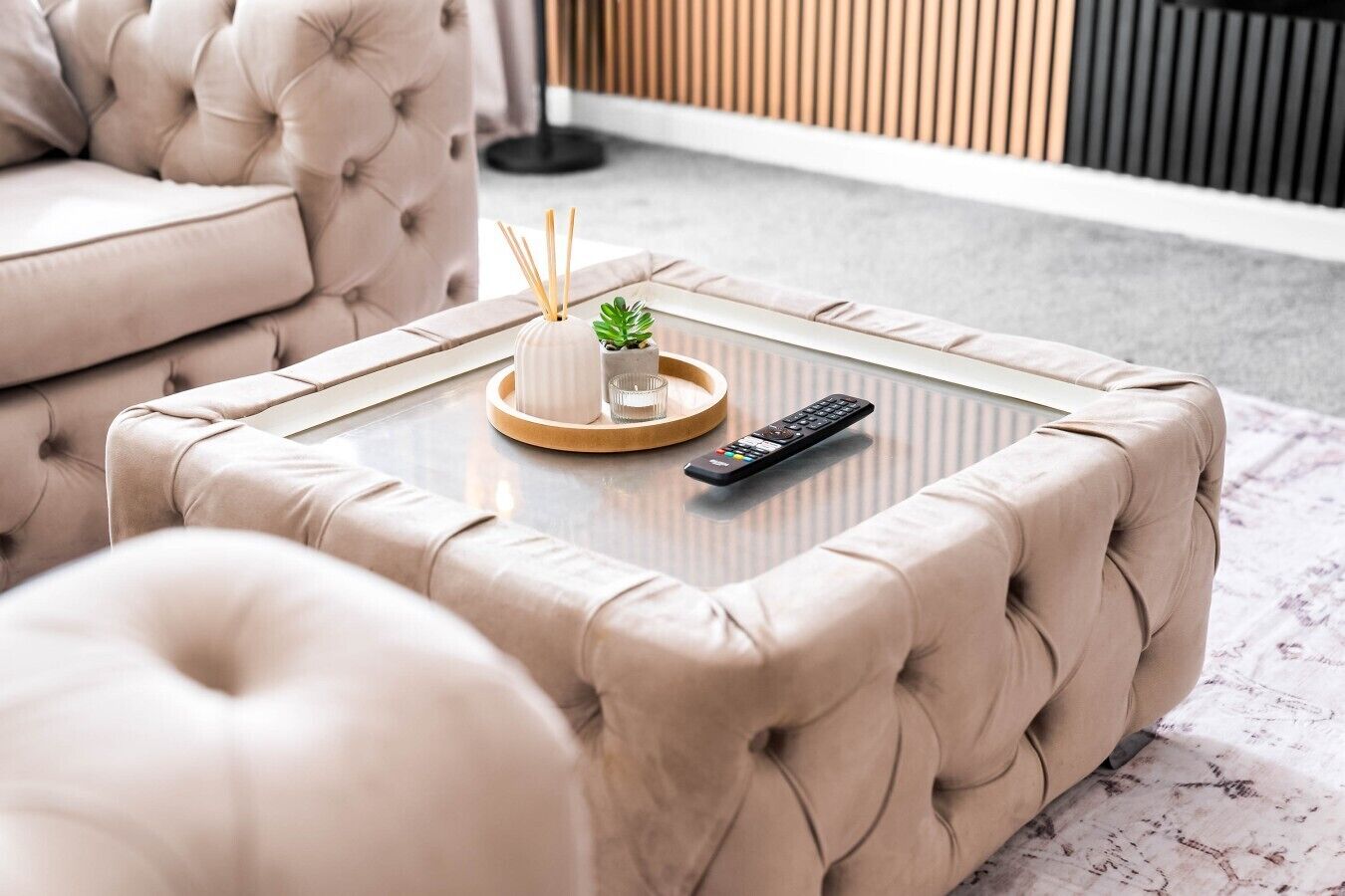 Ein modernes Wohnzimmer mit einer gepolsterten Couch, einem Glastisch mit Fernbedienung und einer dekorativen Ablage mit Pflanze und Kerzen.