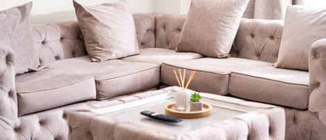 A beige sectional sofa with tufted detailing and matching cushions, a small ottoman with a remote control, and a tray with a candle and reed diffuser.