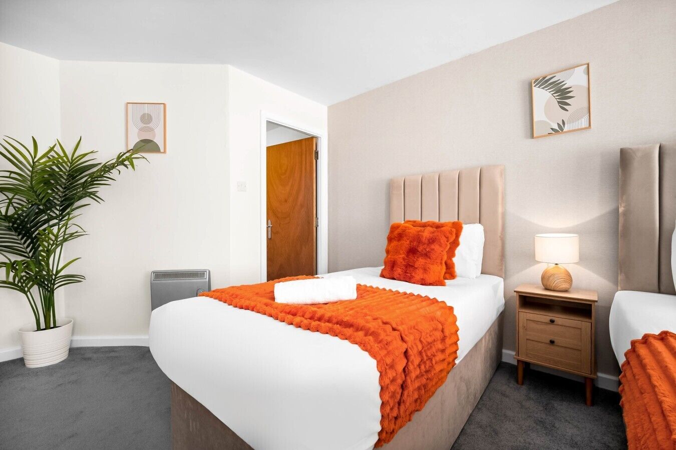 Une chambre d’hôtel avec un lit, une table de chevet avec une lampe, une plante en pot et une porte donnant sur une autre pièce.