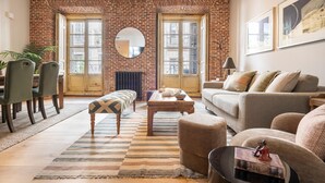 A living room with a brick wall, large windows, a round mirror, a sofa, a coffee table, and a dining area with chairs. - Waou Justicia VI - Short-Term Rental (Madrid)