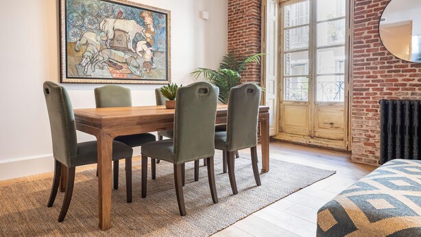 A dining area with a wooden table and chairs, a large painting on the wall, and a view through a glass door. - Waou Justicia VI - Short-Term Rental (Madrid)