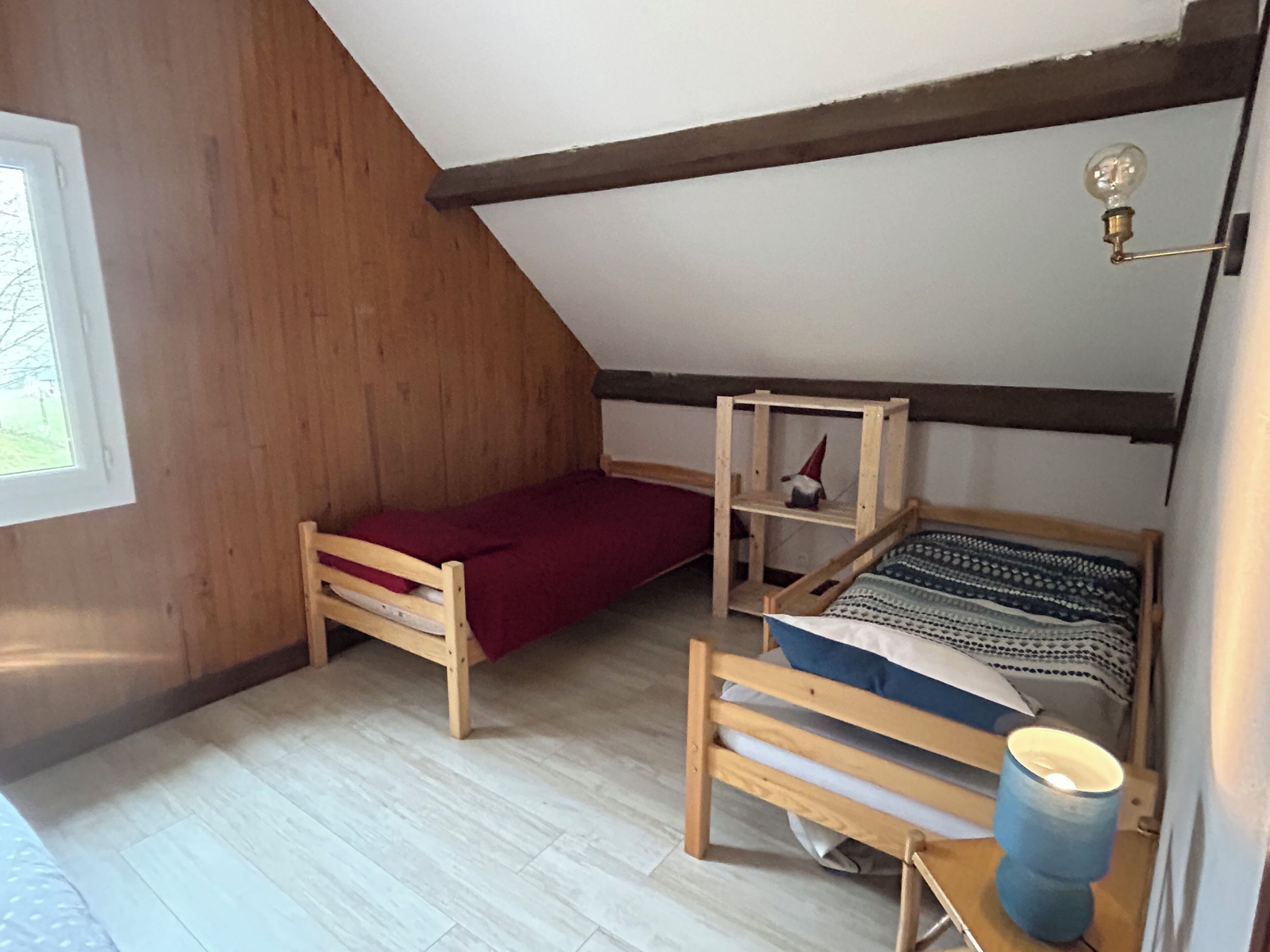 Una habitación pequeña y acogedora con techo inclinado, paredes de madera, una cama individual con un edredón estampado, un estante con un objeto decorativo y una ventana con vistas a la vegetación.