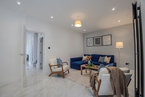 A modern living room with a blue sofa, a white armchair, and a wooden coffee table. There are framed artworks on the wall and a floor lamp.