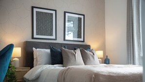 A neatly made bed with a white and beige duvet, a dark headboard, and multiple pillows in shades of blue, gray, and beige. There are two framed pictures on the wall above the bed, and a bedside lamp is visible on the left. - Spacious 5Bed House in Ruislip Sleeps 10 (Ruislip)
