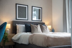 Ein ordentlich bezogenes Bett mit einer weißen und beigen Bettdecke, einem dunklen Kopfteil und mehreren Kissen in Blau-, Grau- und Beigetönen. An der Wand über dem Bett hängen zwei gerahmte Bilder, und auf der linken Seite ist eine Nachttischlampe zu sehen.