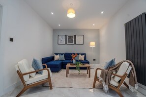 Ein modernes Wohnzimmer mit einem blauen Sofa, zwei Holzstühlen, einem Couchtisch und einer Stehlampe.