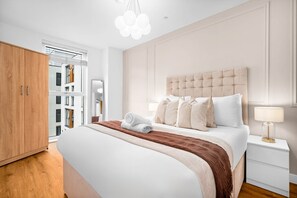 Ein modernes Schlafzimmer mit einem großen Bett, einem Holzschrank, einem Nachttisch mit Lampe und Blick auf ein Gebäude durch ein Fenster.