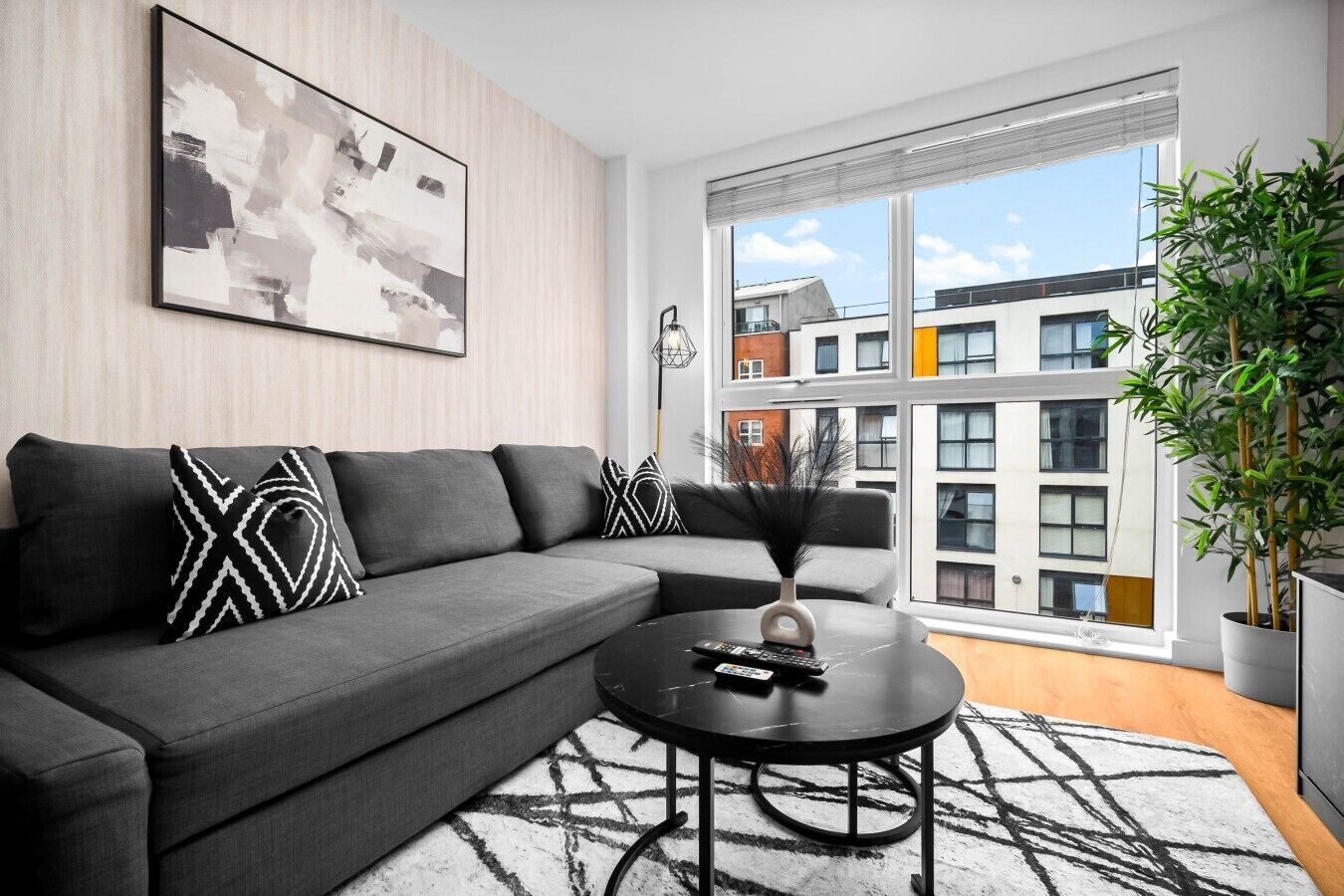 Ein modernes Wohnzimmer mit einem grauen Ecksofa, einem schwarzen Couchtisch und einem großen Fenster mit Blick auf Gebäude.