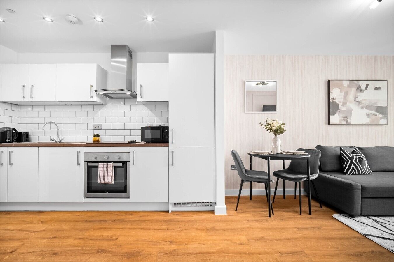 Una cocina moderna con armarios blancos, horno integrado y comedor con sofá negro y una mesa.
