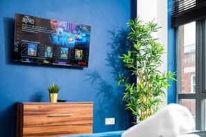 A modern room with a TV displaying a film selection, a wooden dresser, a plant, and a window with blinds. - Luxury 2 Bed Apartment - Parking - JQ - City Centre - Bedroom TV (West Midlands)