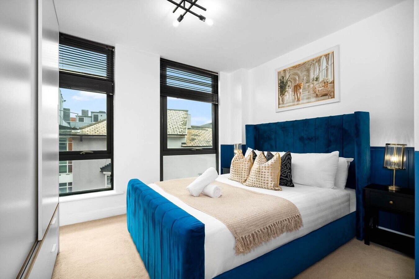 Un dormitorio moderno con una cama grande, dos ventanas, un cuadro y una mesita de noche.