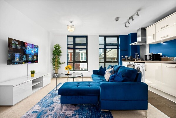 A modern living room with a blue sofa, a flat-screen TV, a small round table with a plant, and a large window. - Luxury 2 Bed Apartment - Parking - JQ - City Centre - Bedroom TV (West Midlands)
