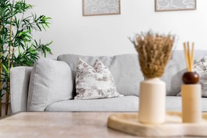 Ein Wohnzimmer mit grauem Sofa, gemusterten Kissen, einer Vase mit Trockenpflanzen und einem Holztisch.