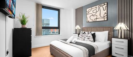 A modern hotel room with a large bed, a flat-screen TV, a potted plant, a window with a view, and a decorative wall piece.