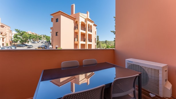 A balcony with a glass table and chairs, an air conditioning unit, and a view of a building with balconies. - Baia da Luz Resort | 2BR with Pool by LovelyStay (Praia da Luz)