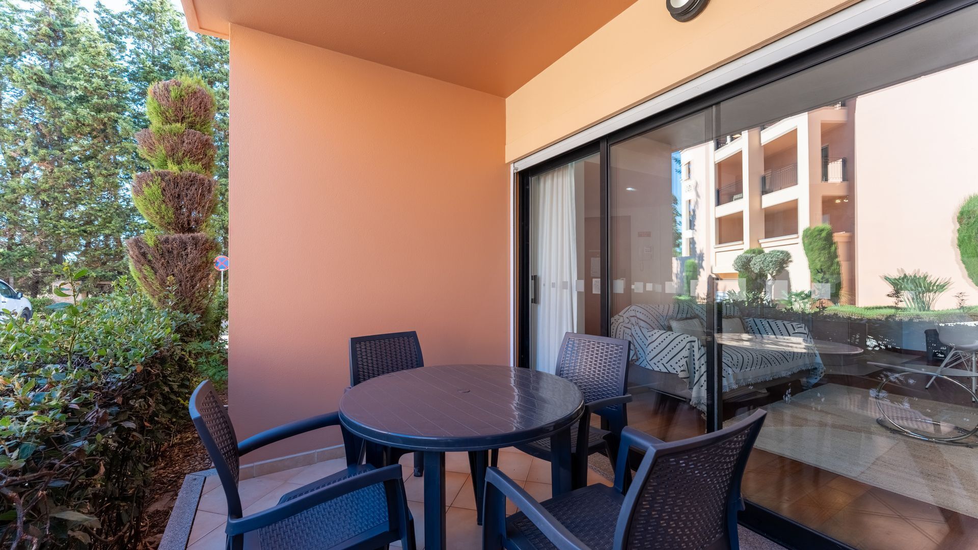 A balcony with a round table and chairs, a sofa, and a glass door leading to an indoor area with a view of a building.