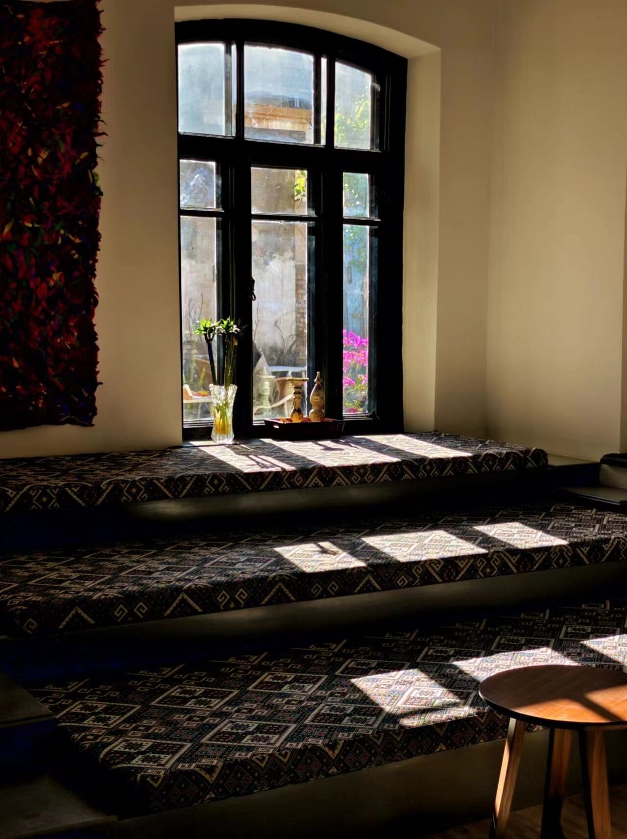Una habitación con un ventanal grande, una alfombra estampada, una mesita y un adorno en el alféizar.