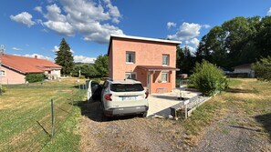 Um bairro residencial com uma SUV estacionada, uma casa com telhado vermelho e um céu claro.