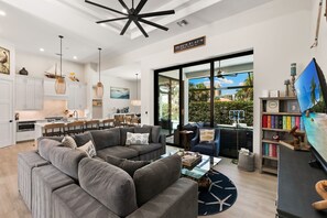 Ein modernes Wohnzimmer mit grauem Ecksofa, einem Essbereich mit Tisch und Stühlen, einer Küche mit weißen Schränken und einem großen Fenster mit Blick ins Freie.