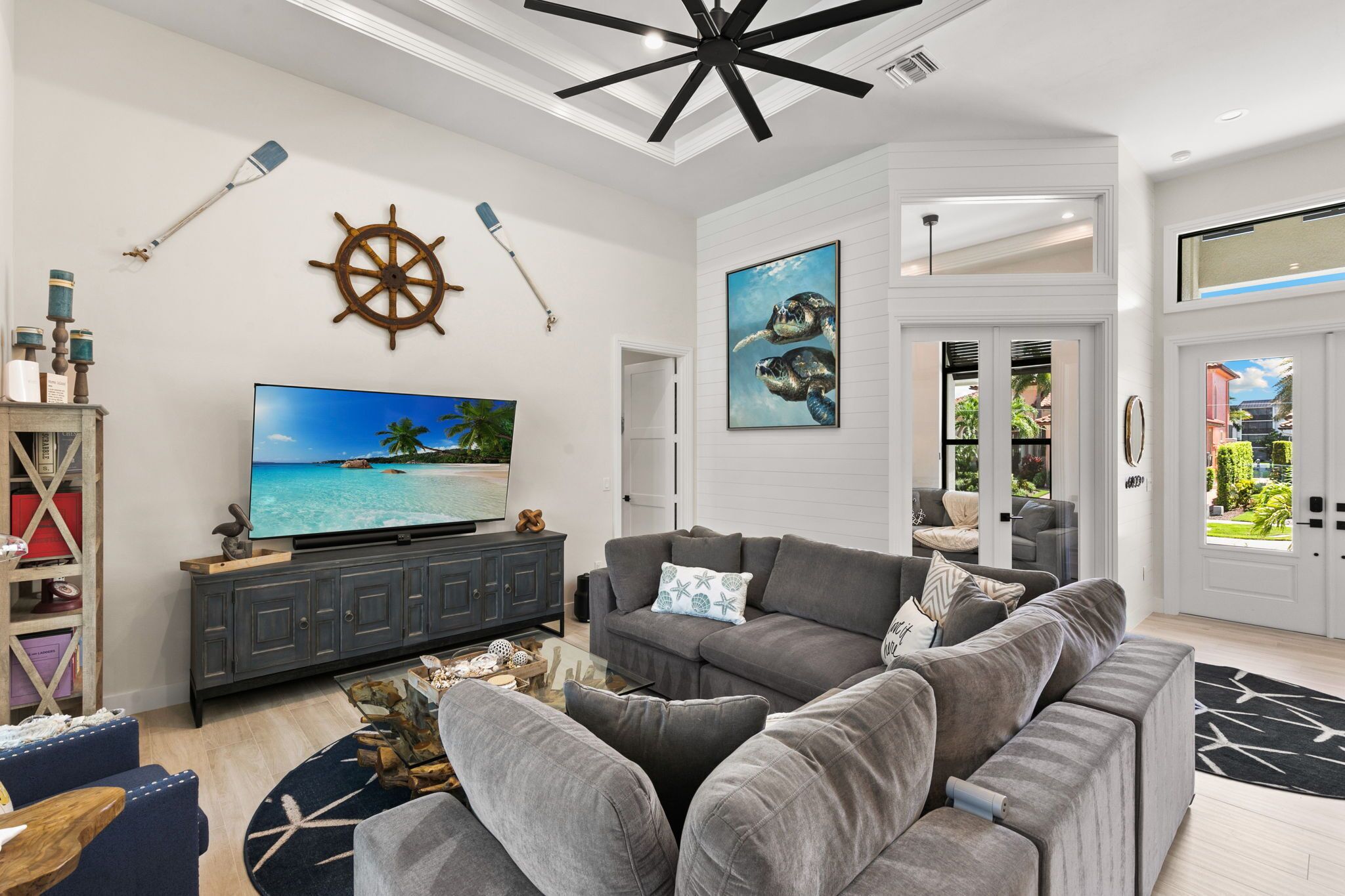 A modern living room with a grey sofa, a flat-screen TV, and nautical decor including a ship's wheel and a painting of sea turtles.