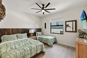 Ein Schlafzimmer mit zwei Betten, einem Deckenventilator, einem Nachttisch, einer Lampe, einer Kommode und einem Fenster mit Blick.